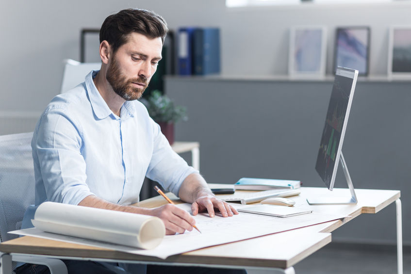 Young male architect, designer making drawings and project on whatman paper on desk in his office. Sitting at a table, with a computer, focused